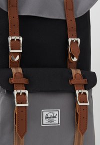 Sac à dos gris et noir avec des sangles en cuir marron et des boucles argentées, portant une étiquette rectangulaire de la marque Herschel sur la poche avant.