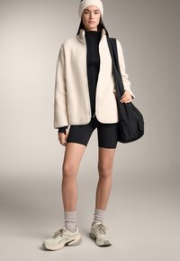 Roomkleurig fleecejack met ritssluiting aan de voorkant, zwart bodypak, zwarte korte broek, grijze sportsokken en lichte sneakers. Zwarte tas over de schouder gehangen.