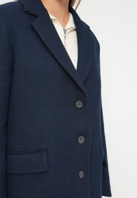 Donna che indossa un cappotto di lana blu navy con tre bottoni sopra un maglione bianco con zip, fotografata dal petto ai fianchi.