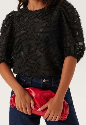 Kobieta ubrana w czarną, teksturowaną bluzkę z bufkami i ciemne jeansy, trzymająca czerwoną skórzaną kopertówkę na wysokości talii.