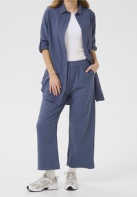Mujer vistiendo pantalones sueltos azules y una camisa a juego con botones sobre una camiseta blanca, con zapatillas y calcetas blancas, de pie con una mano en el bolsillo.
