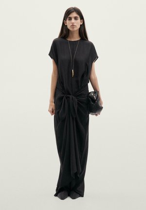 Sort, flydende maxikjole med korte ærmer, knudedetalje ved taljen og glat tekstur. Fremhævet med en guldfarvet vedhængshalskæde og en flettet clutch.