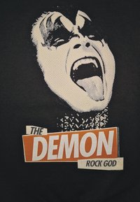 T-shirt nera con una grafica di una persona che urla, con viso dipinto di bianco e grigio, e testo che recita "IL DIO DEL ROCK DEMONIO" in arancione.
