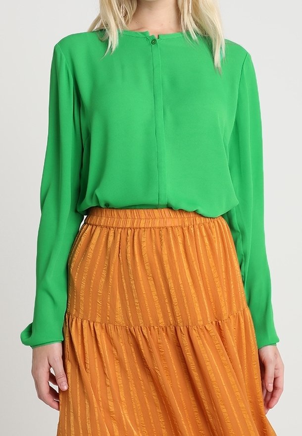 Femme portant un chemisier vert vif à manches longues rentré dans une jupe orange taille haute avec des rayures verticales texturées.