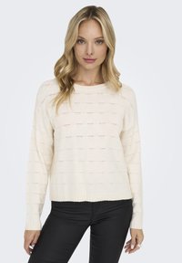 Licht crèmekleurig sweater met een getextureerd patroon en ribborstels. Lange mouwen, ronde hals en een losse pasvorm. Draagt met zwarte, strakke broeken.