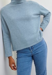 Světlá modrá pletená roláková svetr s volným střihem, dlouhými rukávy a mírně zkráceným lemem, doplněná modrými džínami.