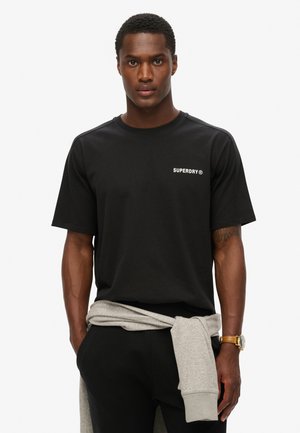 Zwarte katoenen t-shirt met korte mouwen, voorzien van een klein wit "SUPERDRY" logo op de linkerborst. Draagt een grijze trui die rond de taille is geknoopt.
