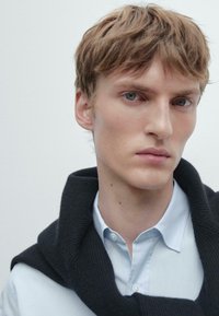 Světle modrá košile na knoflíky s tmavým pleteným svetrem položeným kolem krku, který má hladkou texturu a čistý design.