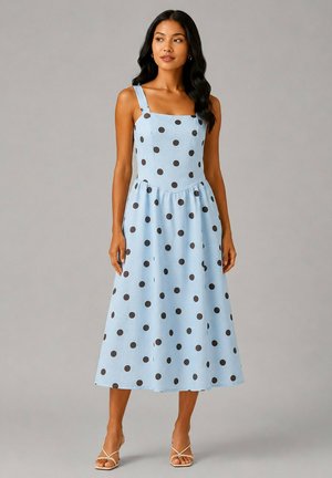 Femme portant une robe midi sans manches bleu clair à pois noirs avec un décolleté carré, associée à des sandales à lanières beiges, debout sur un fond gris.