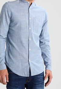 Hellblaues Hemd mit Stehkragen, langem Ärmel und Brusttasche; aus strukturiertem Stoff mit einem subtilen Muster.