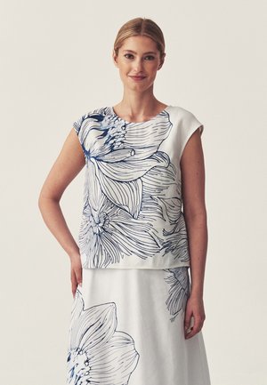 Mujer que lleva una camiseta blanca sin mangas y falda con grandes dibujos lineales florales en azul marino, de pie con una mano en la cadera.