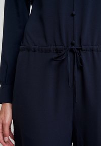 Combinaison bleu marine avec des manches longues, une taille à cordon de serrage et des boutons décoratifs sur le devant. Tissu lisse avec une texture mate.