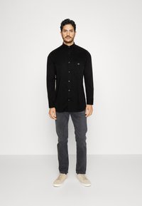 Black long-sleeve button-up shirt with a collar, pocket detail, and a smooth texture, paired with gray jeans and beige sneakers.