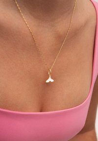 Collier en or avec un petit pendentif en forme de nageoire de baleine, doté d'une finition lisse. La chaîne est délicate, reliée dans un motif en chevrons, sur un haut rose.