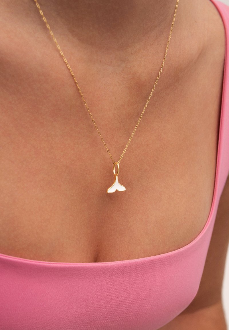 Collier en or avec un petit pendentif en forme de nageoire de baleine, doté d'une finition lisse. La chaîne est délicate, reliée dans un motif en chevrons, sur un haut rose.