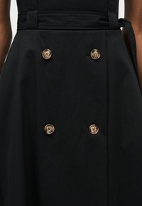 Μαύρο ένδυμα με τέσσερα μεγάλα μπεζ κουμπιά τοποθετημένα σε δύο κάθετες σειρές στην περιοχή της μέσης μπροστά, φορεμένο από ένα άτομο.