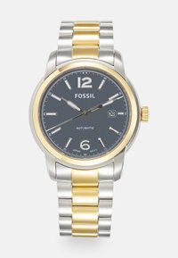Montre automatique Fossil avec un cadran gris foncé, un bracelet en métal argenté et doré, et des index horaires. Présente un affichage de la date et des accents dorés.