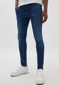 Man som bär mörkblå slim-fit jeans och vita sneakers, står mot en enfärgad ljus bakgrund med händerna i fickorna.