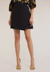 Falda negra envolvente con un detalle de botón en la cintura, hecha de una tela suave. La modelo lleva sandalias amarillas de tiras, que presentan un diseño minimalista.