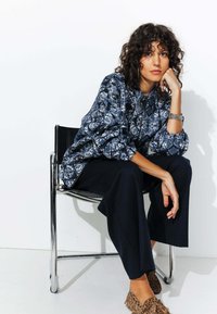 Veste oversize à motifs bleus avec des manches texturées, portée avec un pantalon large noir et des mocassins à imprimé animal. Modèle assis sur une chaise.