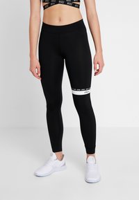 Kvinna som bär svarta tights med vit rand och text på höger lår, vita Nike-sneakers och en svart Nike sport-bh.