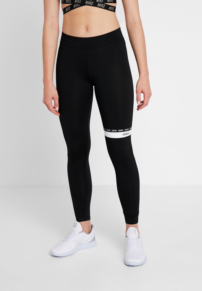 Kvinna som bär svarta tights med vit rand och text på höger lår, vita Nike-sneakers och en svart Nike sport-bh.