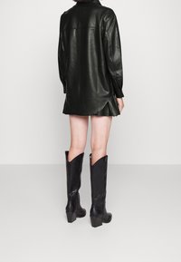 Černé kožené šaty ve stylu košile s křivkovým lemem, dlouhými rukávy a zapínáním na patentky, v kombinaci s černými kozačkami na podpatku po kolena s křivkovým vrškem.