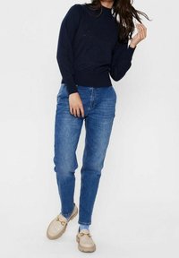 Vrouw in een marineblauwe sweater met lange mouwen, blauwe high-waisted jeans, lichtbeige instappers met gouden details, staand tegen een effen achtergrond.
