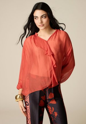 Vrouw met lang donker haar die een doorschijnende koraalblouse met ruches draagt en een zwarte broek met rood bloemmotief, geaccentueerd met gouden armbanden.