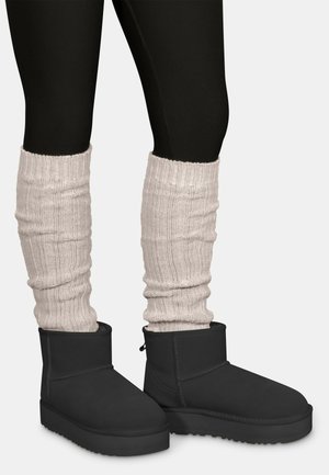 Schwarze Stiefeletten mit flacher Sohle, kombiniert mit texturierten cremefarbenen Stulpen mit Rippenmuster, die über schwarzen Leggings getragen werden.