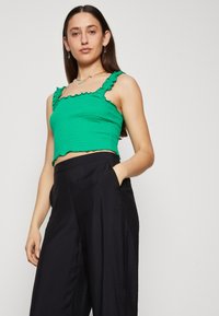 Top court vert avec des bretelles à volants, en tissu texturé, et ourlet en dentelle, associé à un pantalon noir à taille haute et jambes larges.