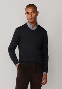 Donkergrijs gebreid sweater met een V-hals ontwerp, gedragen over een lichtblauw geruit shirt, gecombineerd met een paar bruine broeken. Gladde textuur, aansluitende pasvorm.