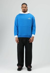 Blauwe hoodie met een witte binnenvoering en zak, gecombineerd met losse zwarte broek en zwarte sneakers met gekleurde veters.