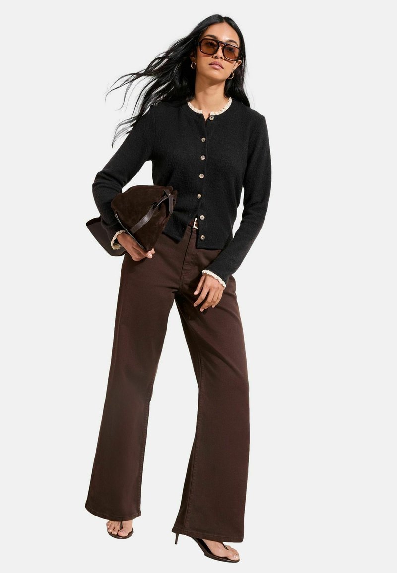 Schwarzer Strickjacke mit Knöpfen und cremefarbenem Besatz, kombiniert mit braunen Hosen mit weitem Bein. Dunkelbraune Wildledertasche und schwarze High Heels vollenden den Look.