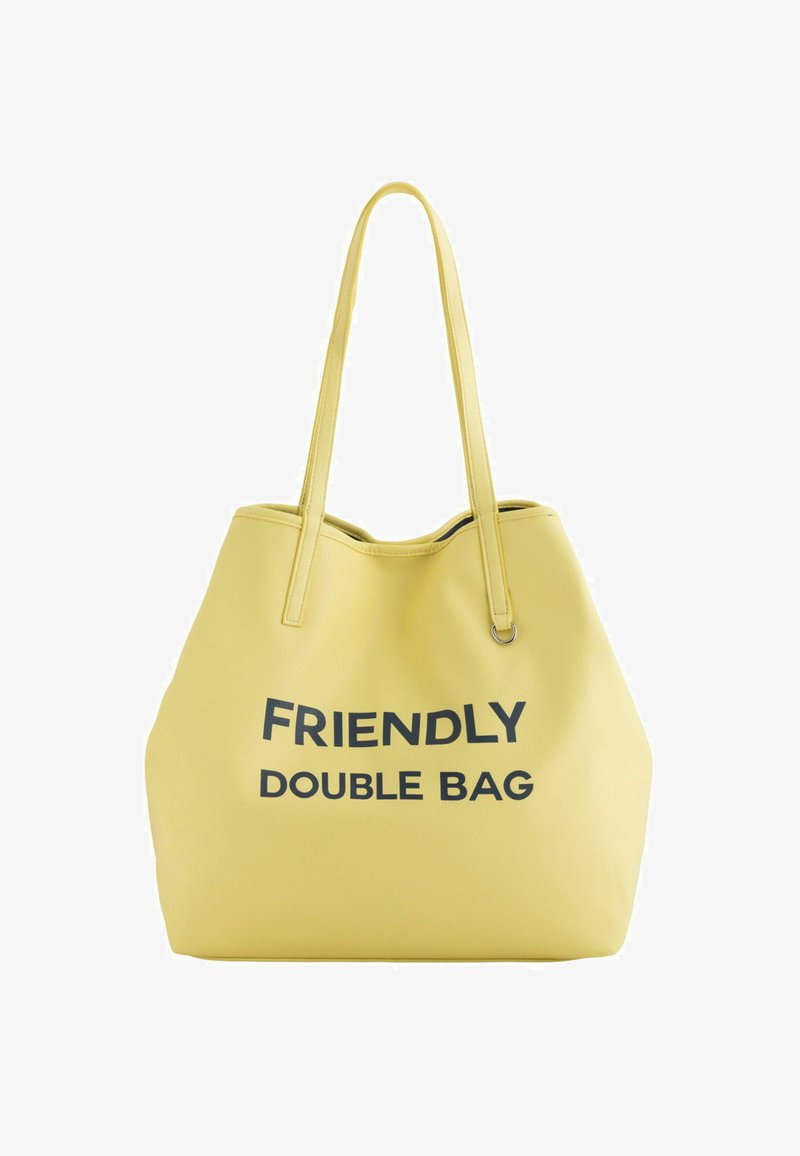 Keltainen kassi, jossa on teksturoidun pinnan, jossa on rohkea musta teksti "YSTÄVÄLLINEN KAKSOISKASSI". Sisältää pitkät, ohuet kahvat kantamista varten.