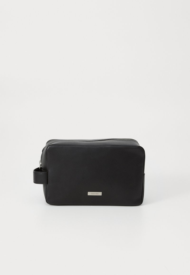 Svart lærtoalettveske med rektangulær form, glidelås, sidehåndtak og sølvfarget logo plate på forsiden. Glatt tekstur.