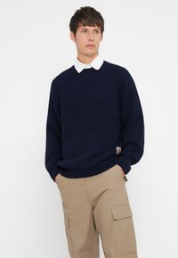 Navy knitted sweater with a ribbed texture, round neckline, and long sleeves, worn over a white collared shirt. Casual beige cargo pants.