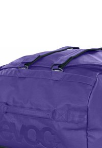 Borsa da viaggio viola con tessuto strutturato, doppi manici superiori, cerniere e dettaglio con logo. Presenta cuciture rinforzate per una maggiore durata.