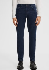 Homme portant un jean slim fit bleu foncé, des bottes noires à lacets et une chemise blanche à manches longues, se tenant devant un fond uni.