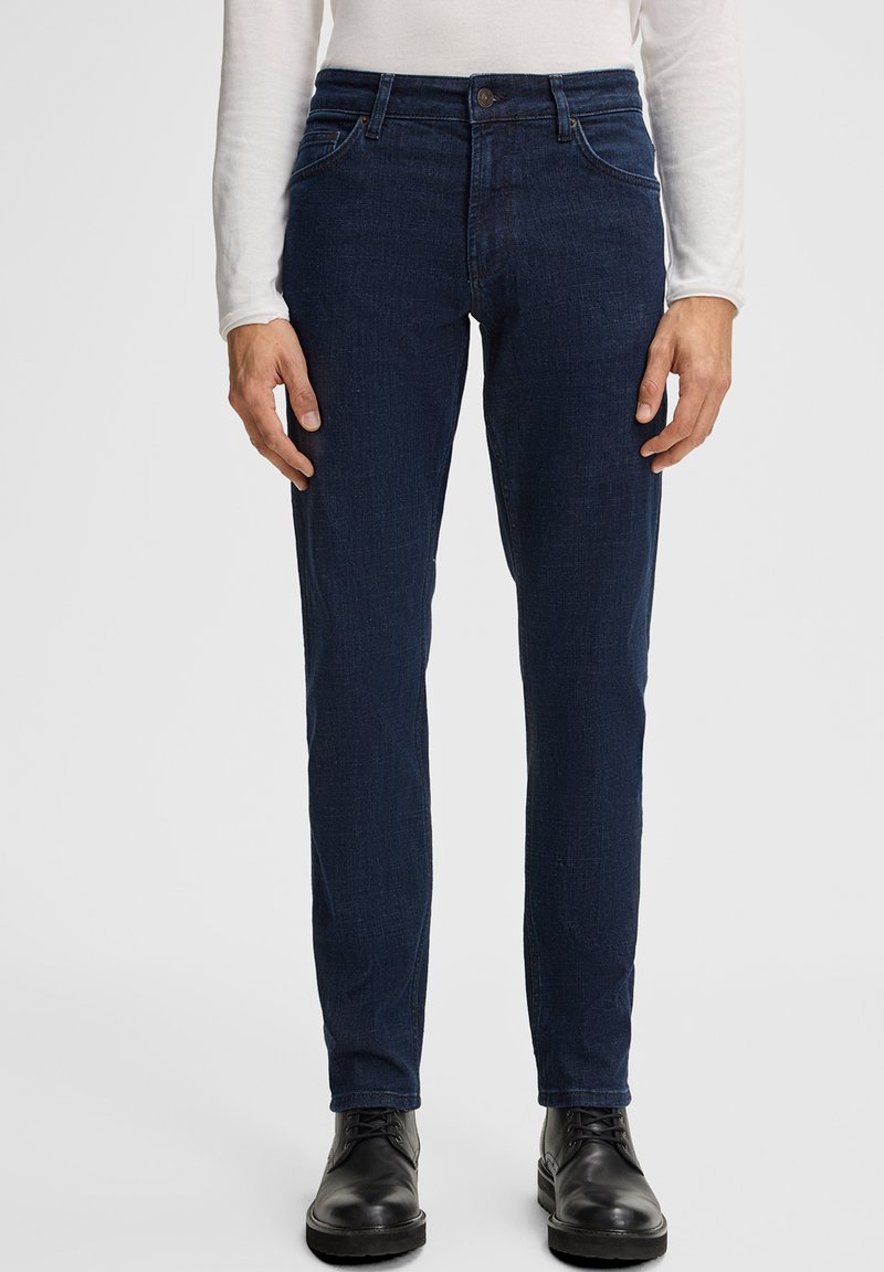 Homme portant un jean slim fit bleu foncé, des bottes noires à lacets et une chemise blanche à manches longues, se tenant devant un fond uni.