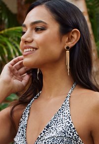 Gold-tone earrings featuring a round black stud and cascading fringe design. The model has dark hair and is wearing a patterned top.