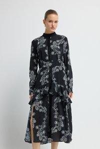 Frau mit zurückgekämmtem Haar trägt ein schwarzes Langarmkleid mit weißem Blumen-Paisley-Muster und gerafftem Mittelteil, steht vor einer weißen Wand.