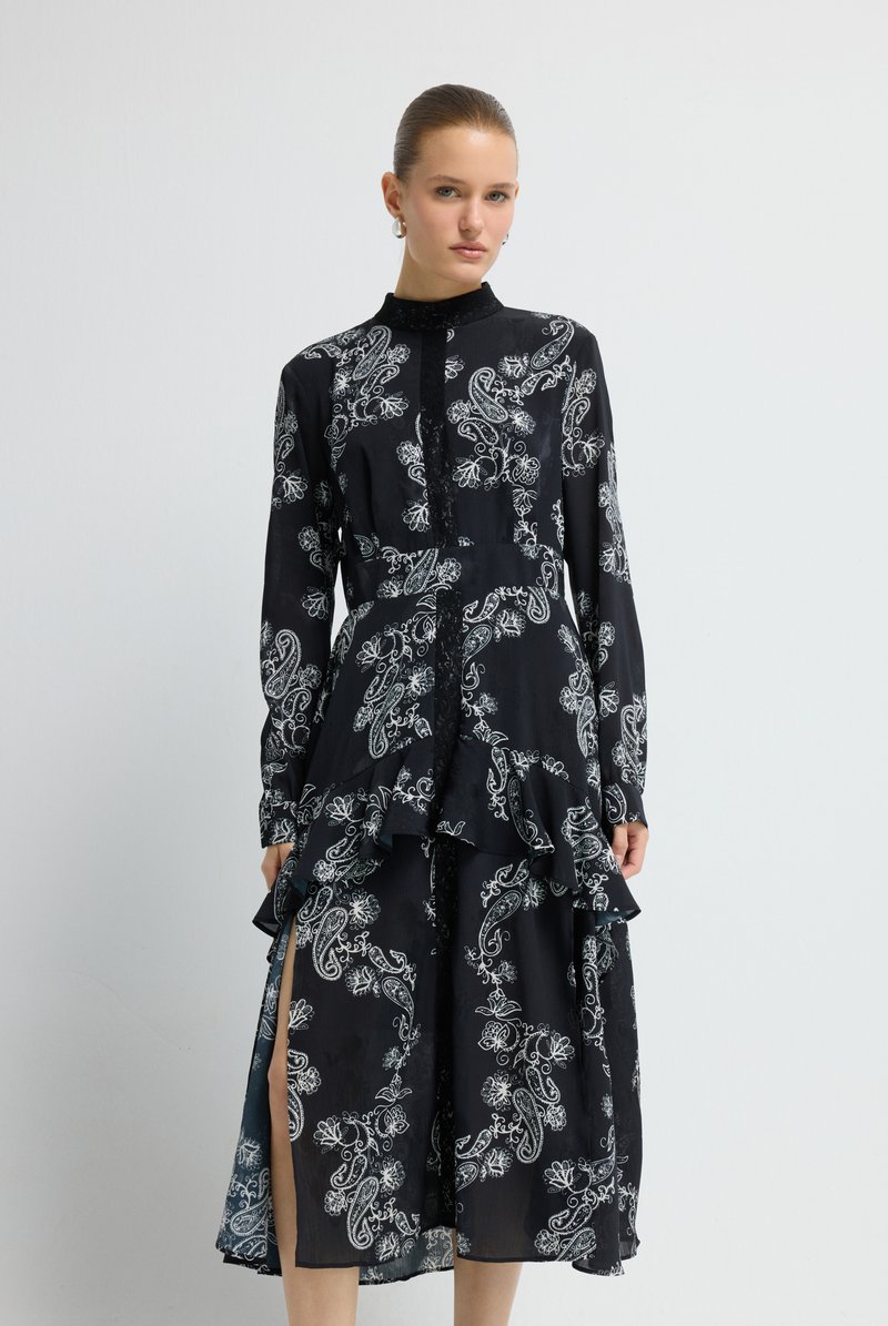 Frau mit zurückgekämmtem Haar trägt ein schwarzes Langarmkleid mit weißem Blumen-Paisley-Muster und gerafftem Mittelteil, steht vor einer weißen Wand.