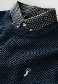 Granatowy sweter ribbed z kraciastym kołnierzykiem. Posiada srebrny emblemat jelenia na lewej piersi oraz teksturowany materiał. Klasyczny dekolt w serek.