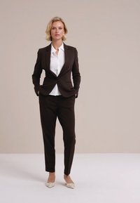 Blondína stojí s rukami v kapsách, oblečená v tmavohnedom kostýme, bielej košeli a bielych špicatých topánkach na jednolatom pozadí.