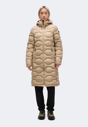 Persoon die een lange beige doorgestikte donsjas met kap draagt, zwarte broek en groene wandelschoenen, staand met het gezicht naar voren.