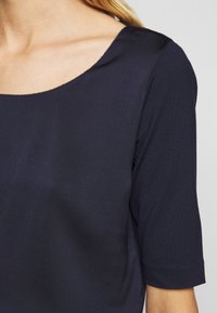 Haut bleu marine en tissu lisse, avec un col rond et des manches courtes ornées de coutures discrètes.