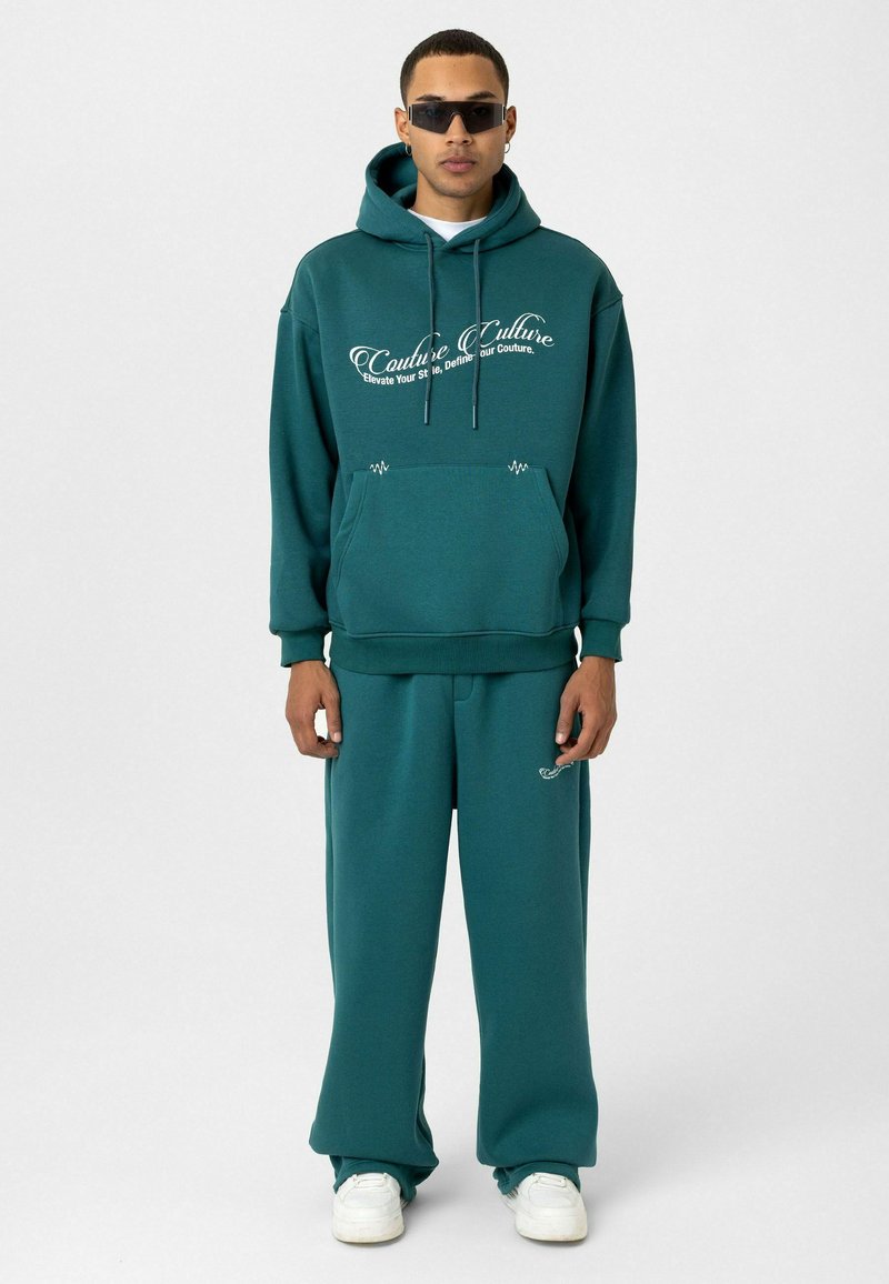Teal hoodie met witte tekst, kangoeroezak en trekkoorden; gecombineerd met bijpassende losse broek en witte sneakers.