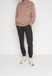 Světle taupe mikina na zip s vysokým límcem a logem. Kombinováno s tmavými tepláky a béžovými teniskami. Měkký bavlněný mix materiálů.