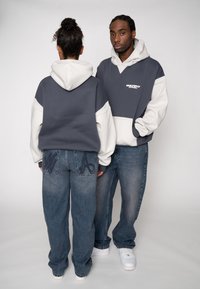 Hanorace gri și albe cu glugă, cu culori contrastante, croială lejeră. Blugi albaștri cu aspect uzat și accente brodate pe buzunare.